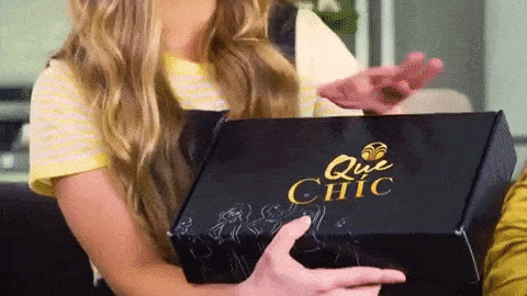a woman holding up a box and she runs her fingers along the top which has the company's name Que Chic on it